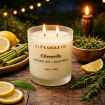 J | F Candle Co. Citronella soy candle with a lit wick in a frosted glass jar, styled outdoors at dusk on a rustic wood surface with lemon slices, lemongrass, and rosemary, with warm ambient lighting in the background