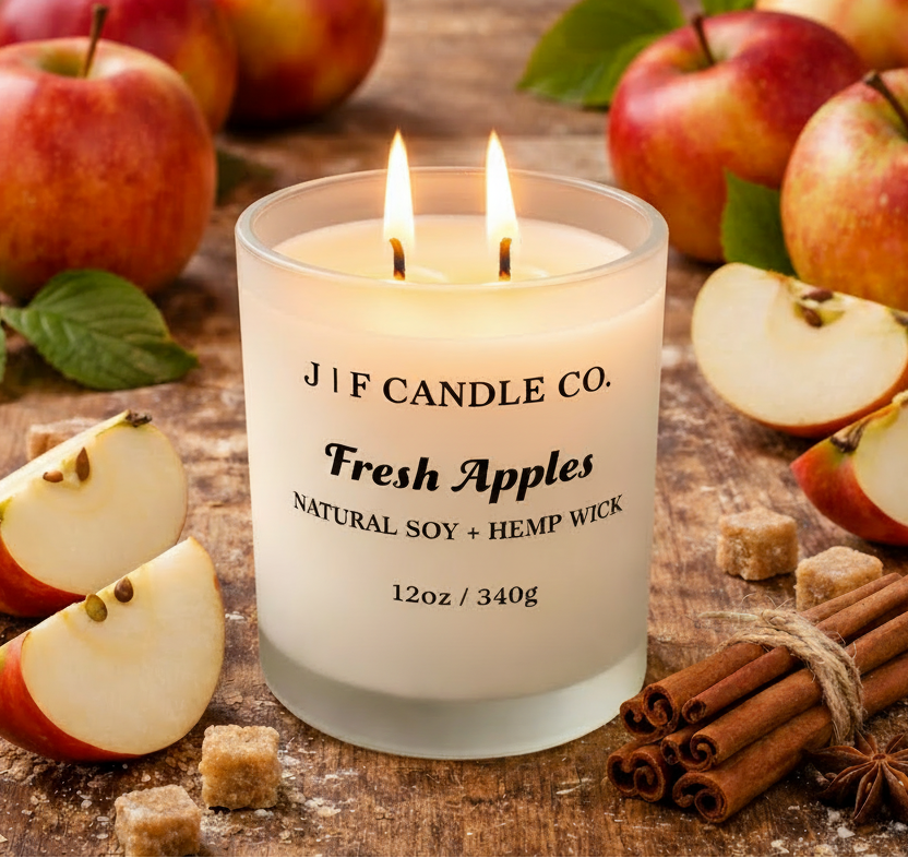 J | F Candle Co. Fresh Apples soy candle with a lit wick in a frosted glass jar, styled on a rustic wood surface with fresh apples, apple slices, cinnamon sticks, and sugar cubes in a warm, natural setting