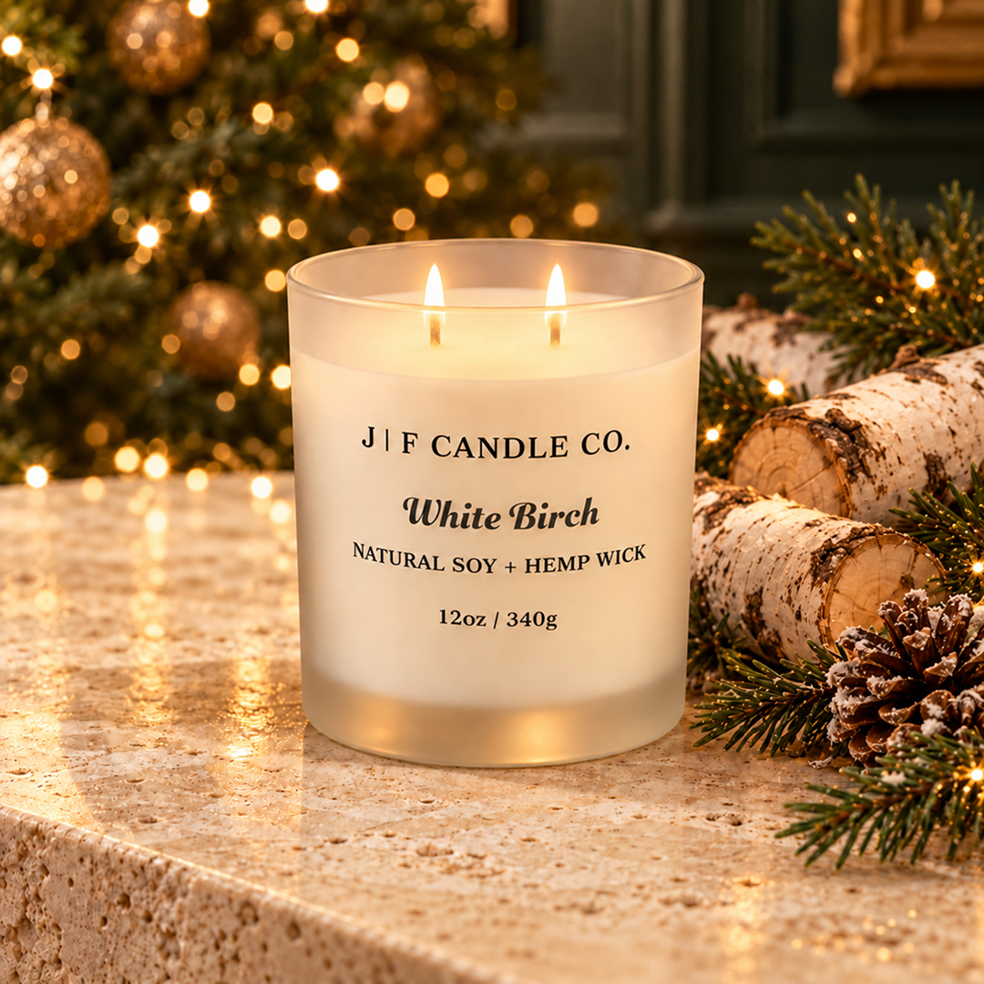 A double-wick scented candle by J|F Candle Co. in a frosted glass jar labeled 'White Birch,' placed on a stone surface with a blurred Christmas tree, lights, birch logs, and pine branches in the background.